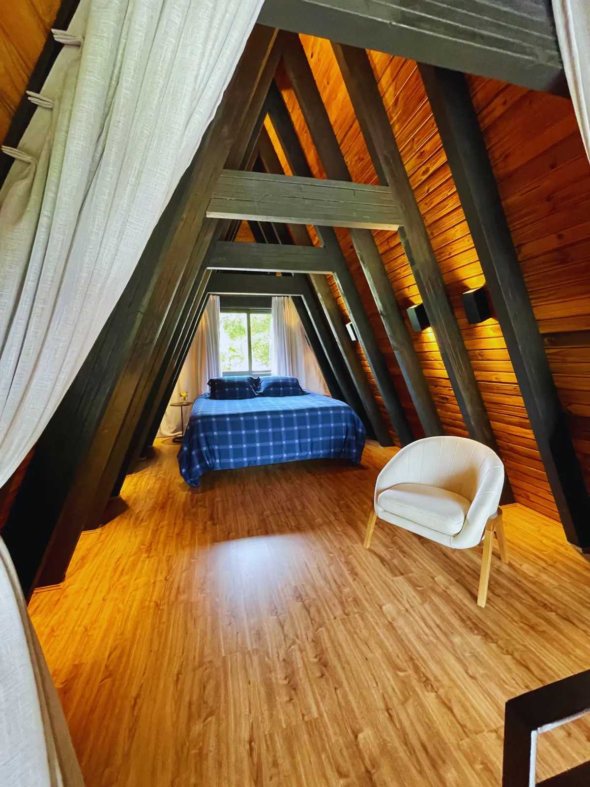Quarto no mezanino sob as vigas do telhado A-frame, com cama king, poltrona e janela para a mata