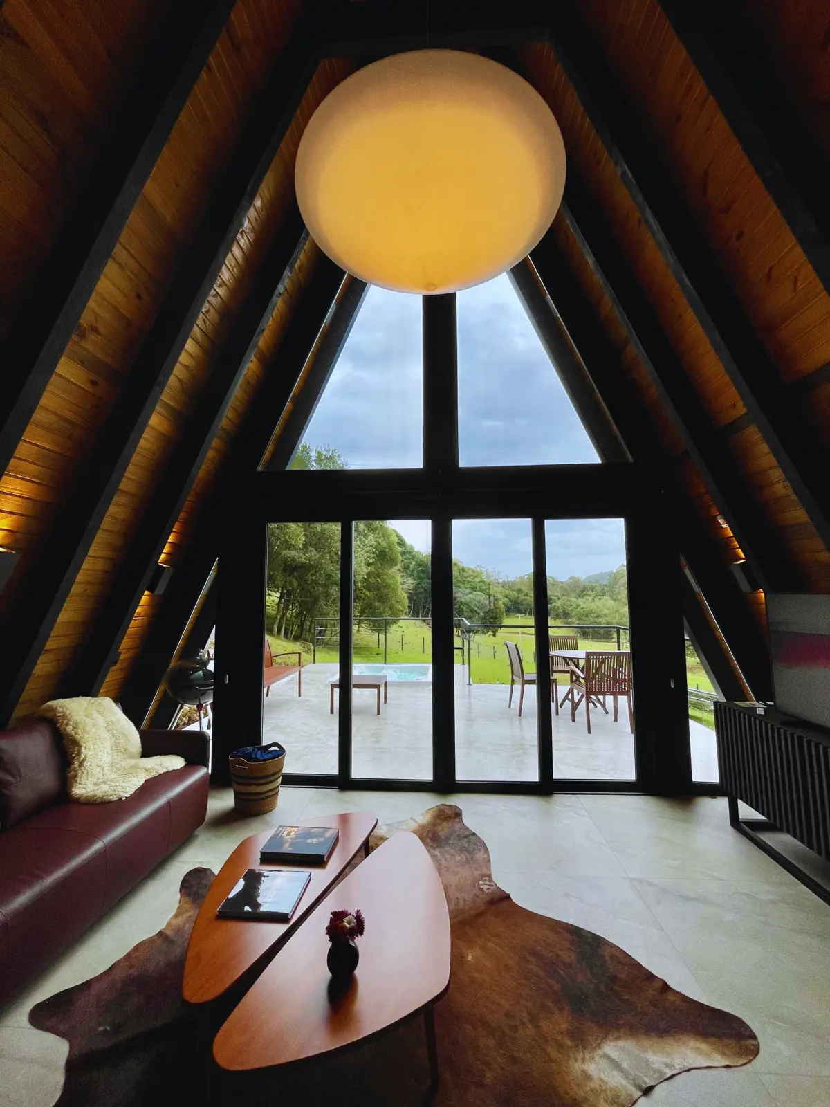 Sala com janelão triangular do A-frame, sofá de couro, lustre pendente e vista para o deck e a mata