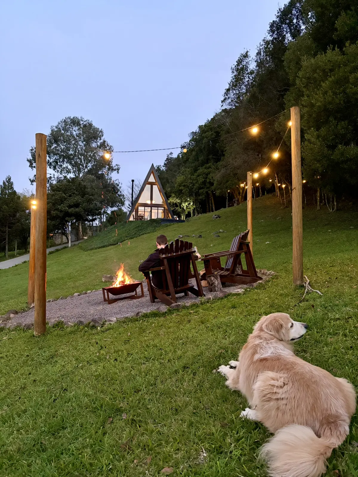 Hóspede junto à fogueira ao entardecer, com golden retriever deitado no gramado
